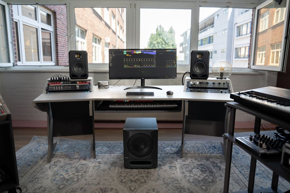 Studio d'enregistrement avec un bureau équipé d'un écran large affichant un logiciel de production musicale, entouré de deux enceintes Adam Audio