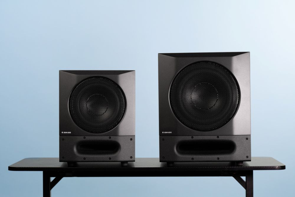 Deux caissons de basses noirs de la marque Adam Audio, de tailles différentes, sont posés côte à côte sur une table devant un fond bleu clair.