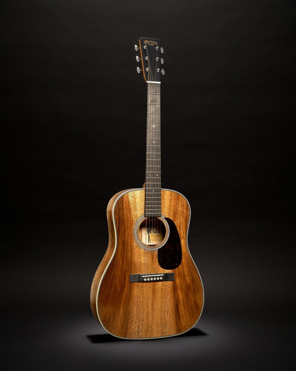 Guitare Martin O’ahu HG-28K2 acoustique à cordes acier avec une table en bois brun aux veines apparentes, vue de trois-quarts sur fond noir.